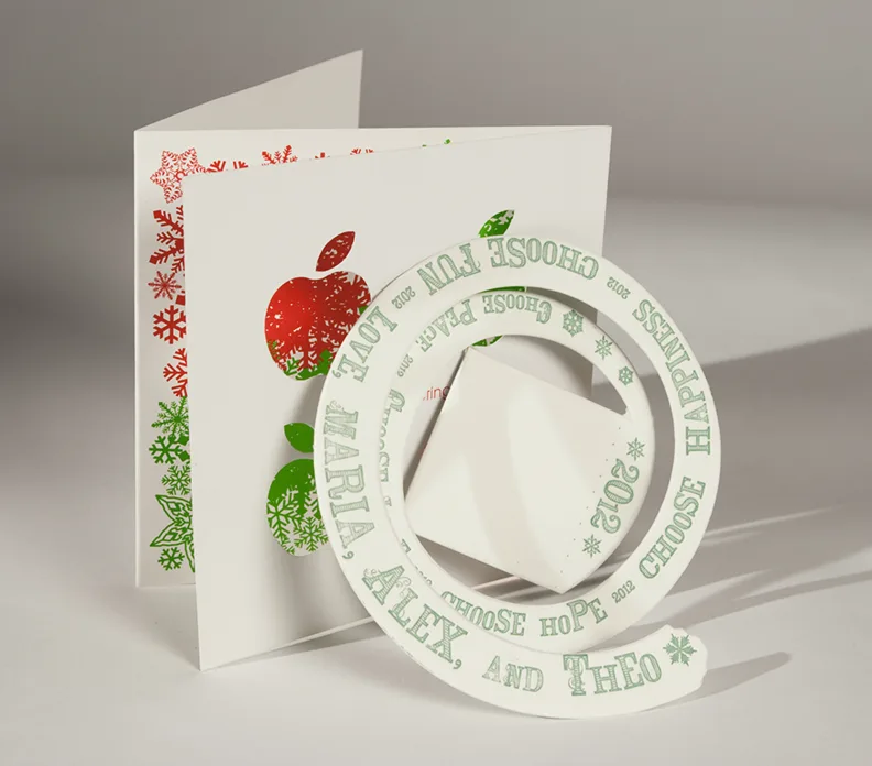 Die-cut holiday greeting card with red apple and green snowflake patterns, circular cutout revealing white envelope inside