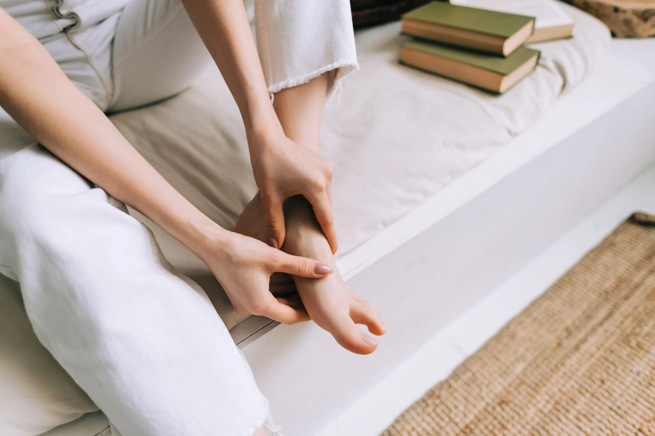 Person massaging their foot while sitting on a bed with books nearby