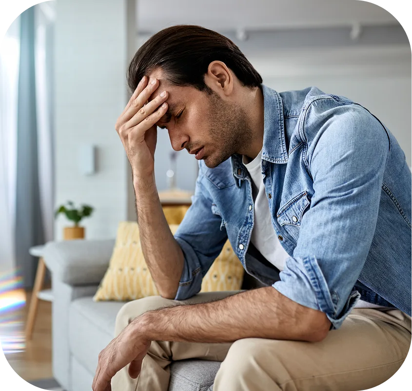 Man in denim shirt sitting on couch with hand to face, appearing distressed