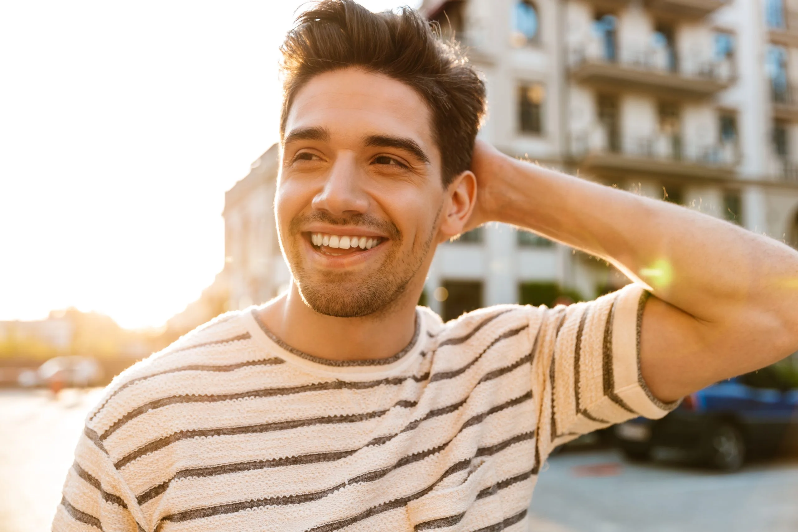 Man in striped shirt smiling outdoors at sunset near apartment building