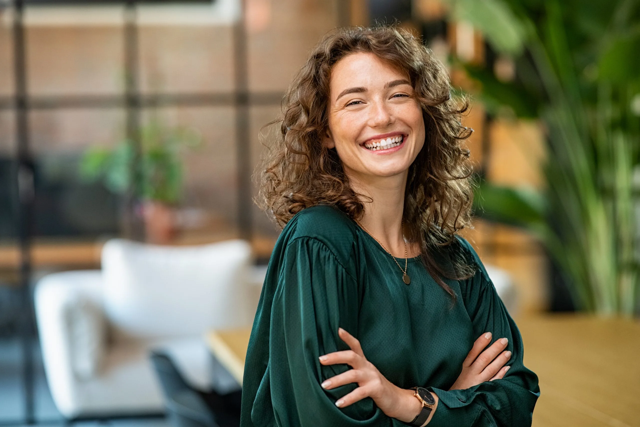 Woman smiling with arms crossed in a bright, plant-filled modern office setting