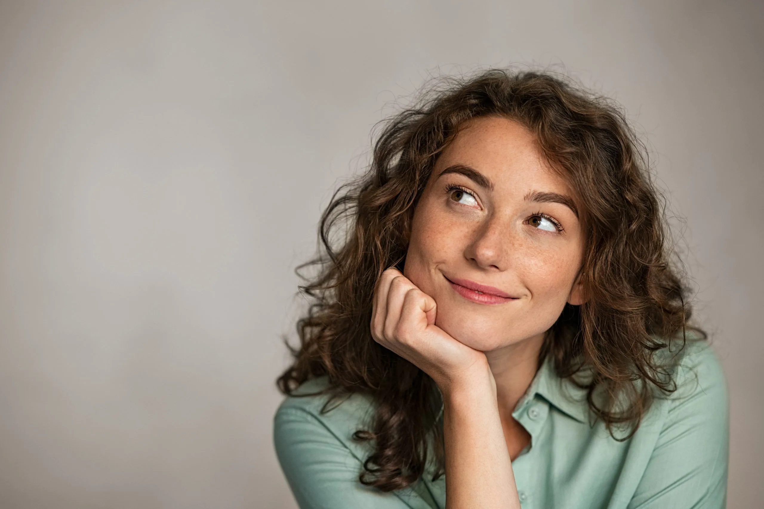 Woman with curly brown hair in teal shirt, hand on chin, smiling while looking upward thoughtfully