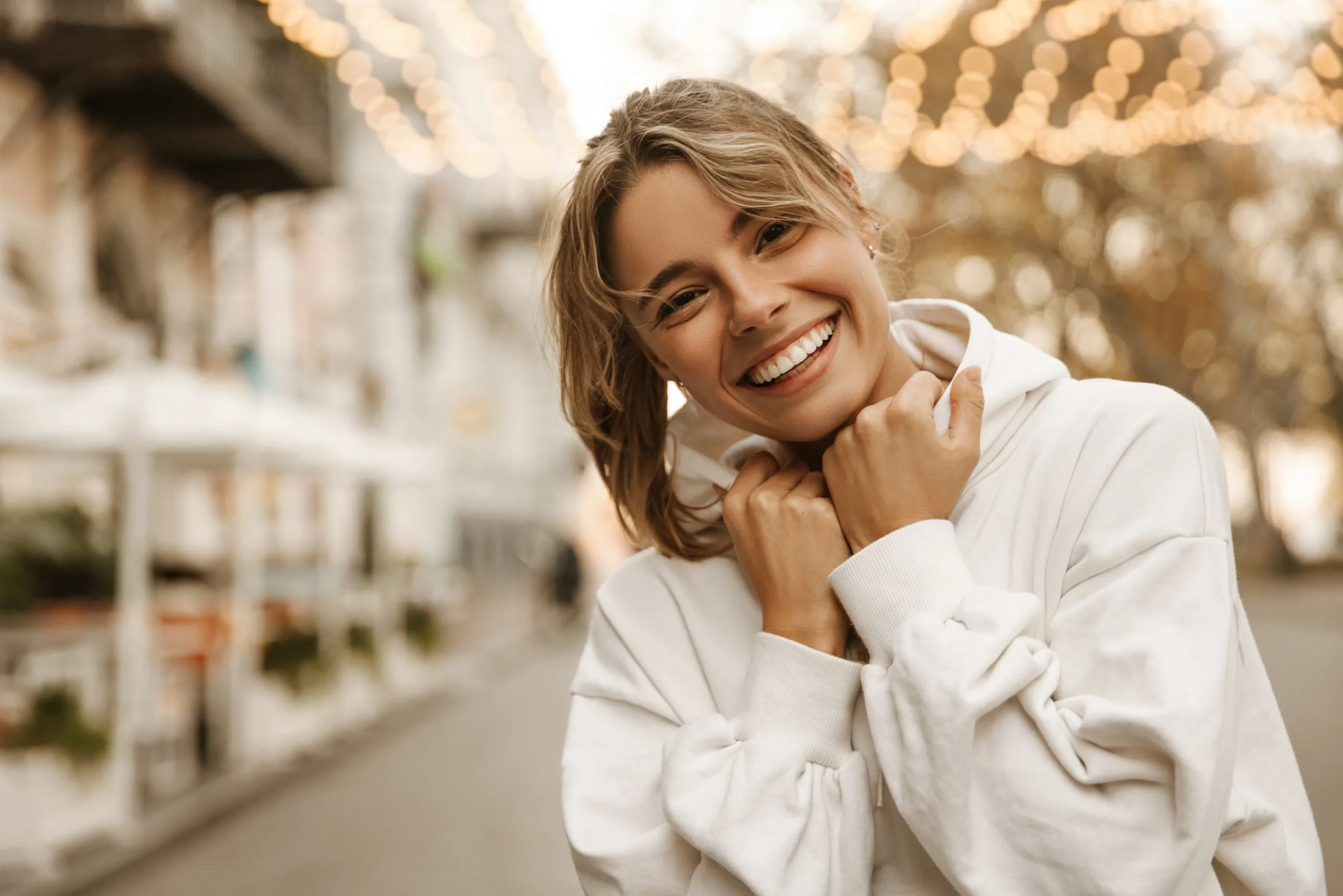 Woman in white shirt smiling outdoors with blurred storefront in background