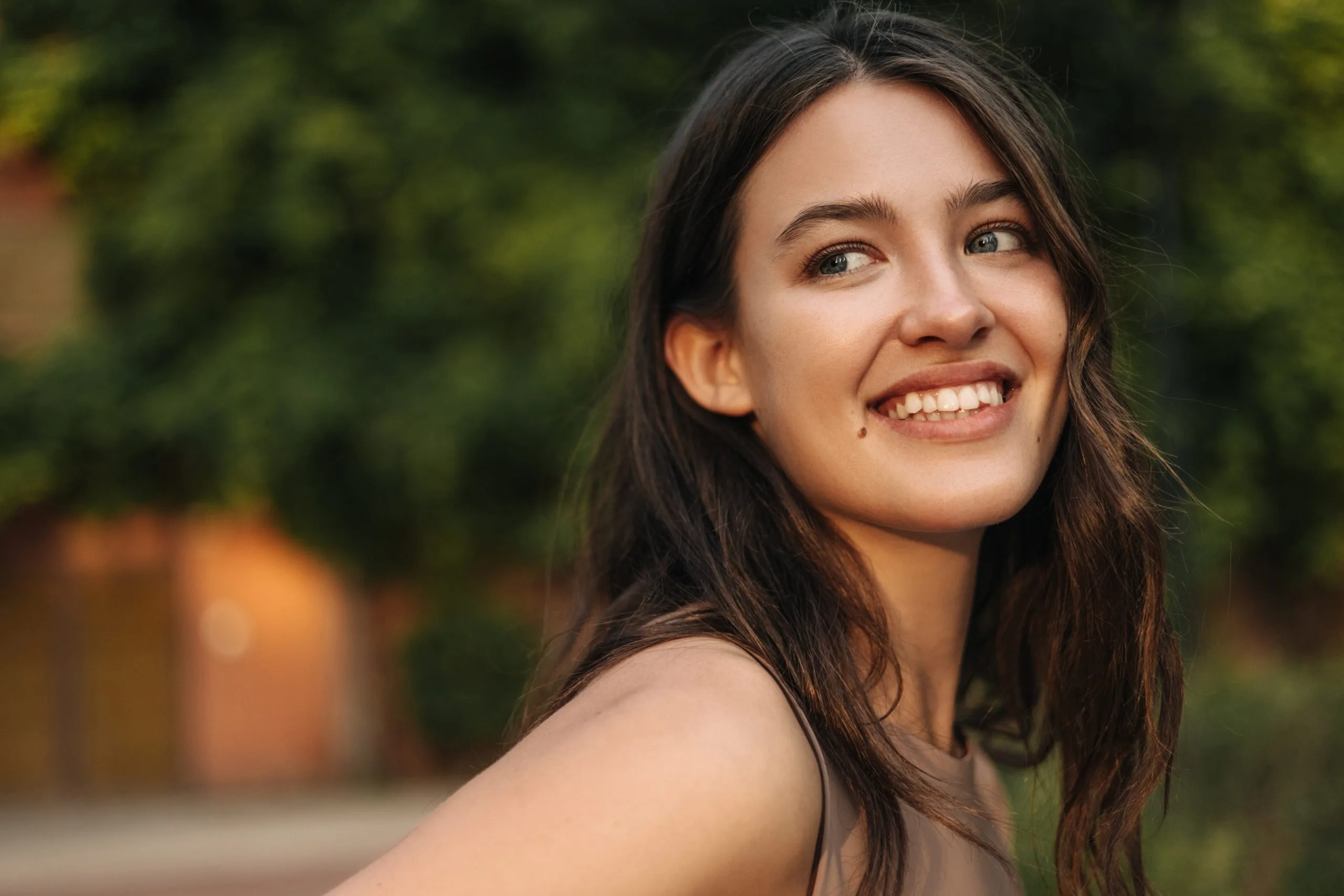 Young woman with long dark hair smiling outdoors with green foliage behind her
