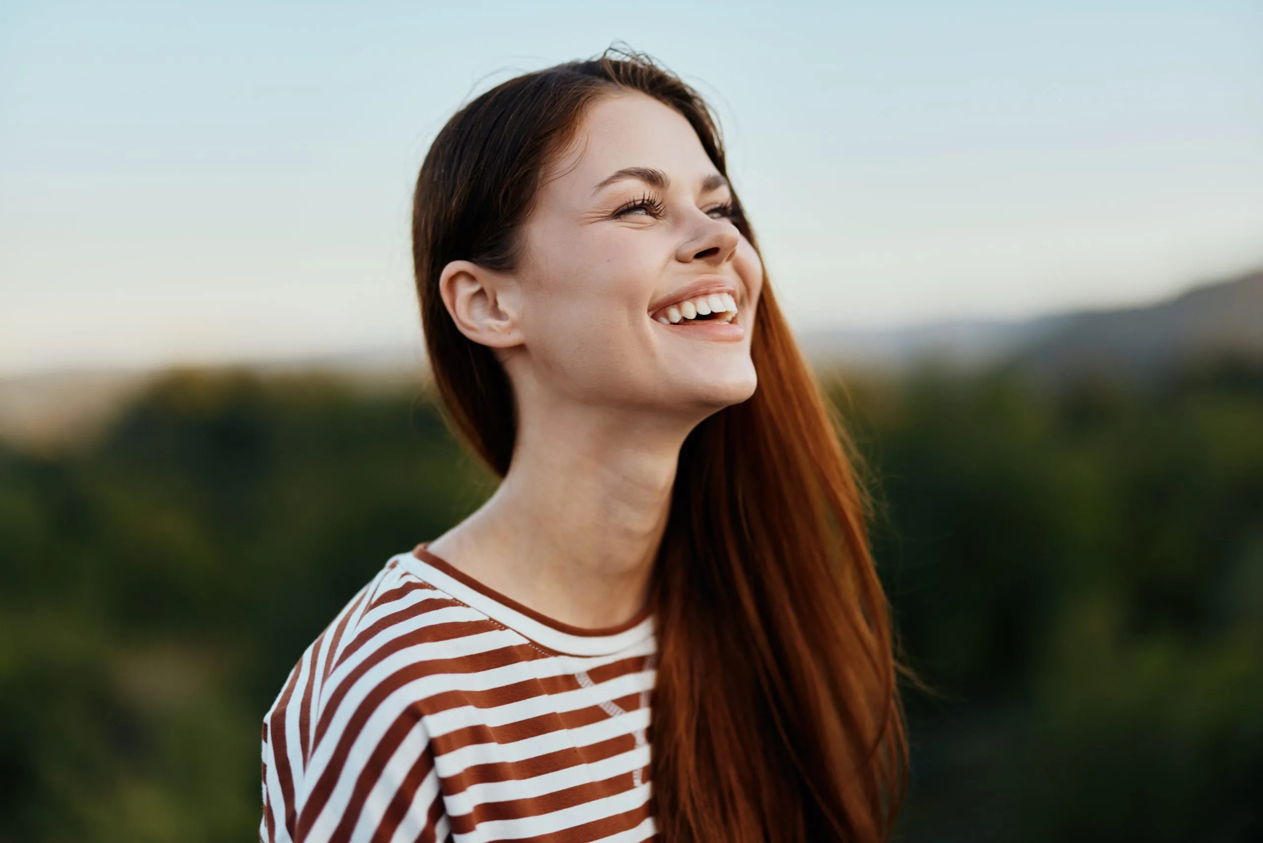 Woman in striped shirt laughing while looking upward outdoors with mountains in background