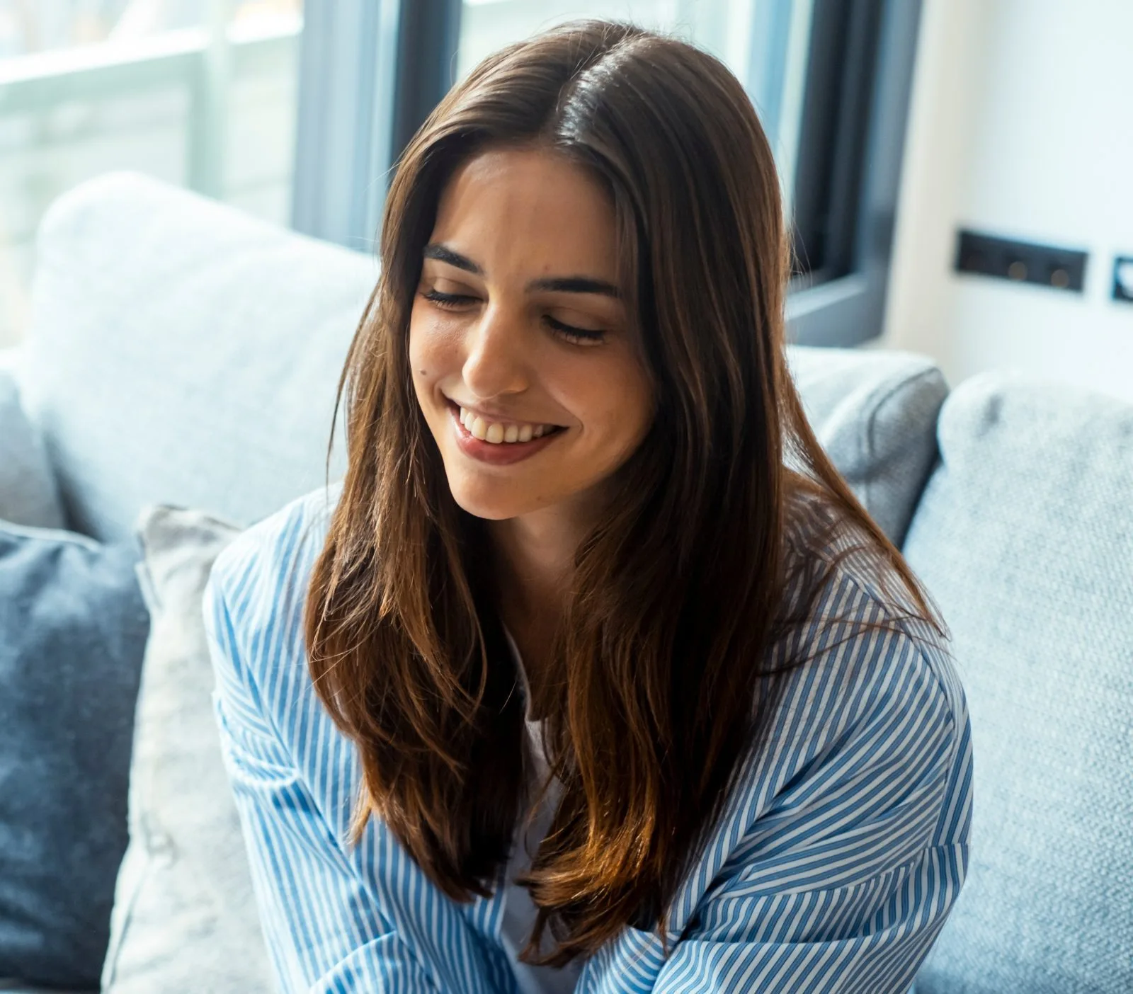 Woman in blue striped shirt smiling while sitting on couch by window