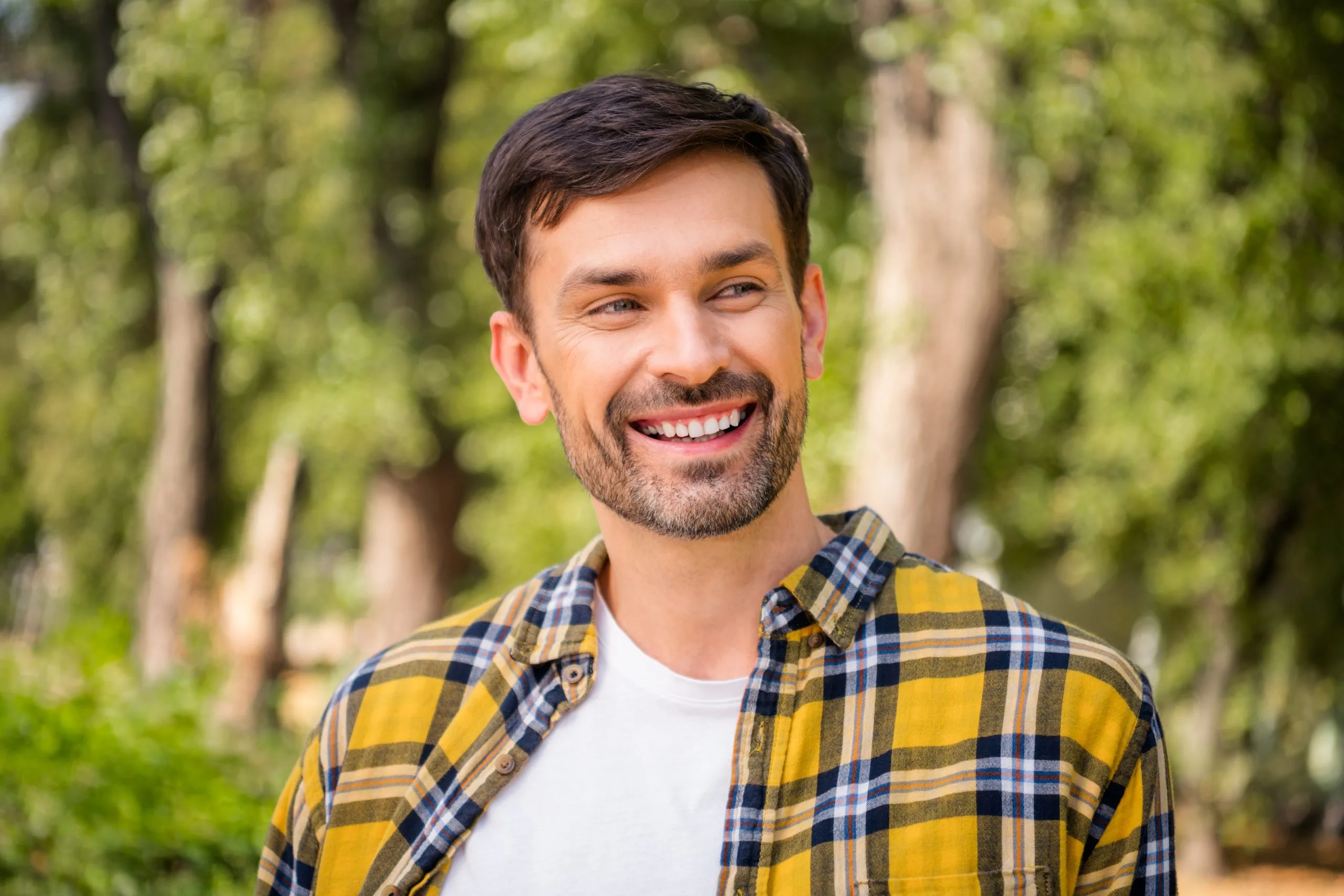 Man smiling outdoors among tall trees, wearing a yellow and navy plaid shirt