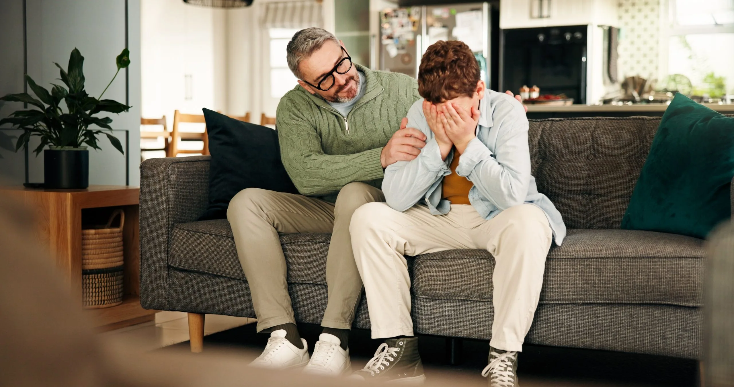 Therapist comforting male patient on couch in home setting, patient has hands to face in distress