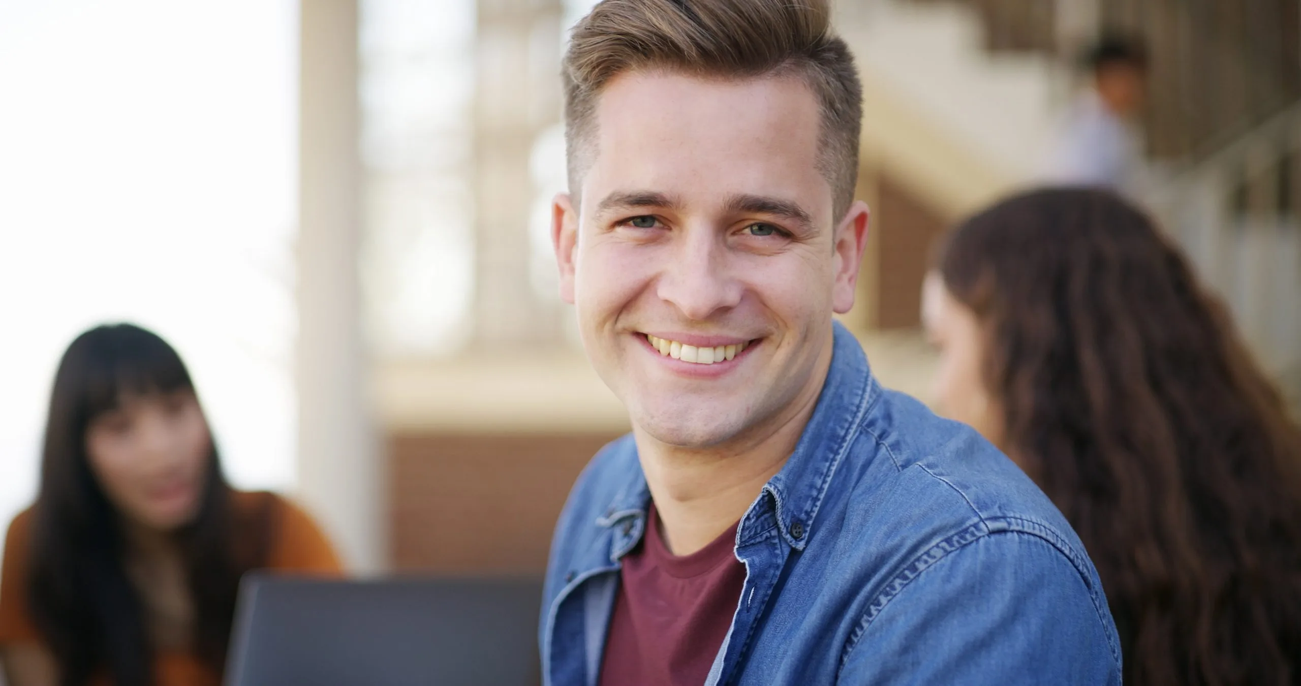 Smiling man in denim shirt with other people blurred in background