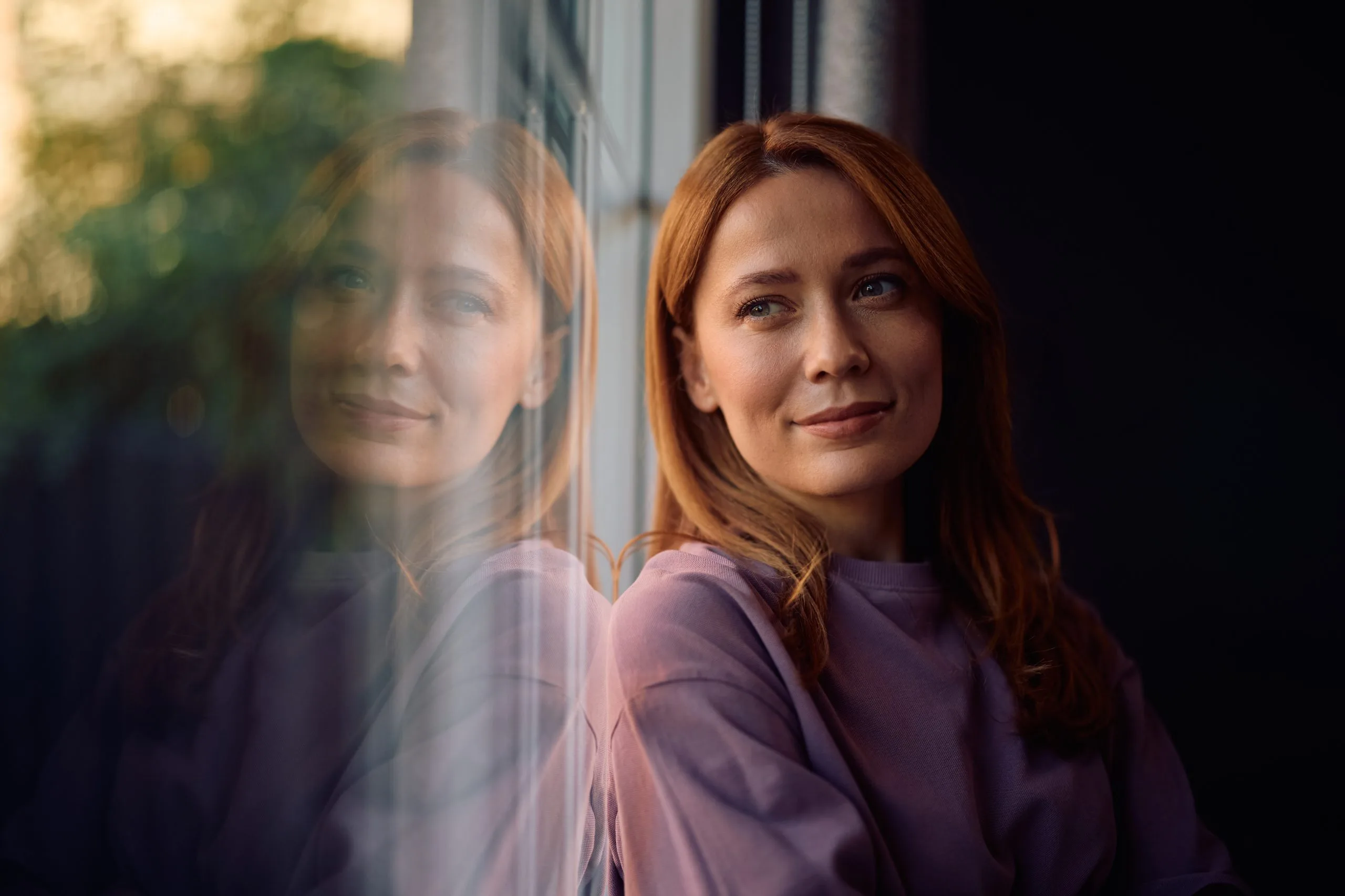 Woman with reddish-brown hair looking out a window with sunlight filtering through