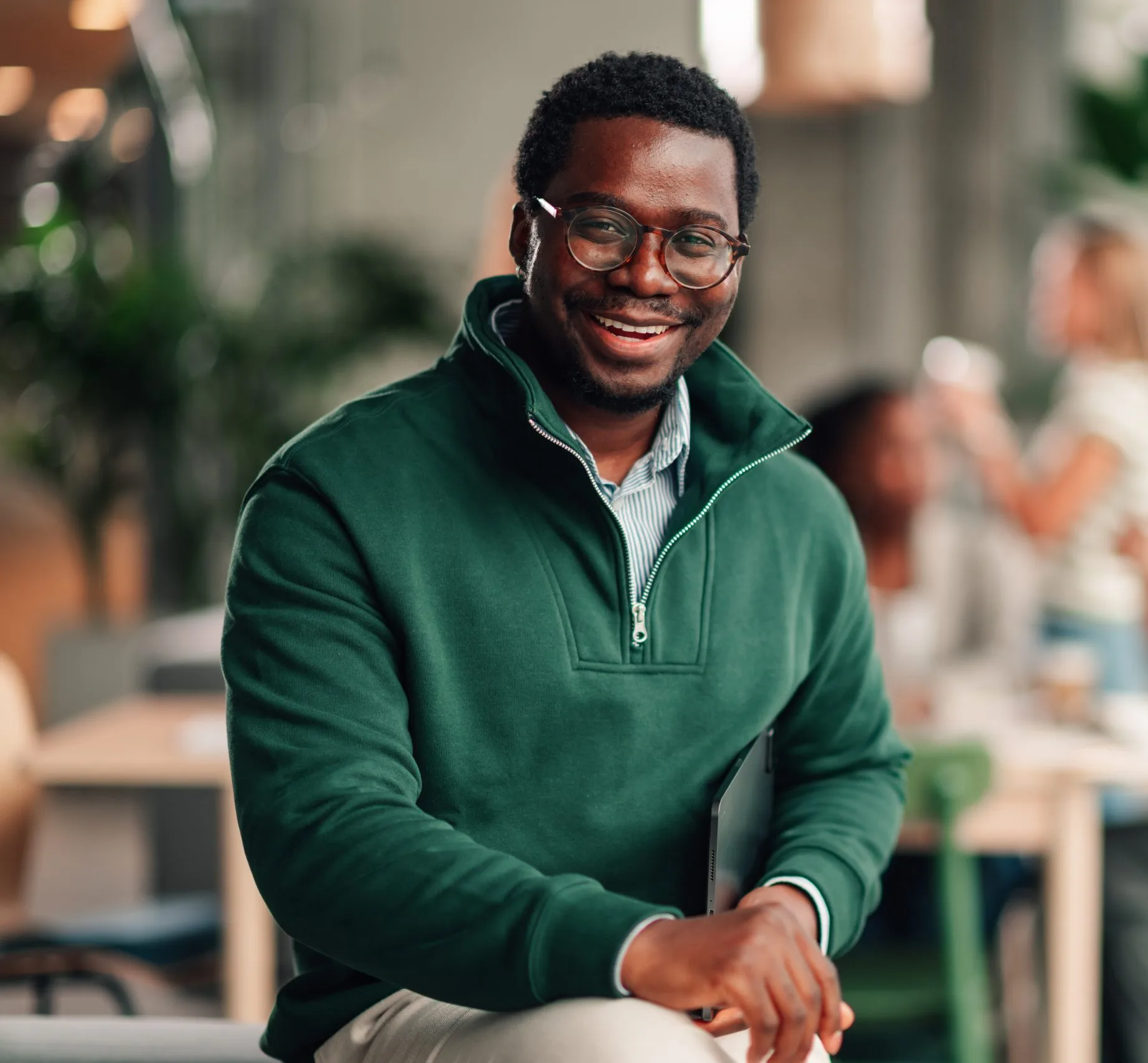 Man smiling while seated in a bright office setting with plants and colleagues in background