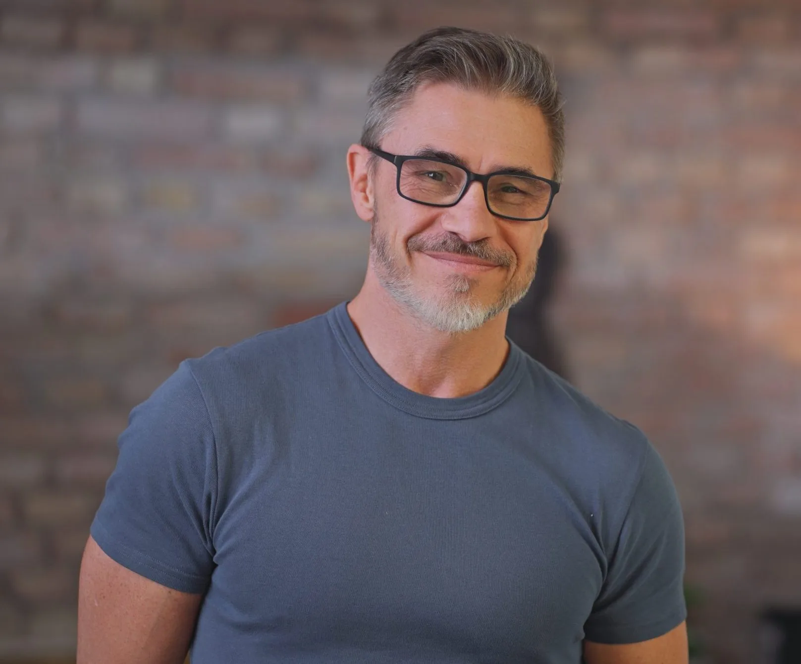 Man wearing glasses and blue t-shirt smiling at camera against brick wall