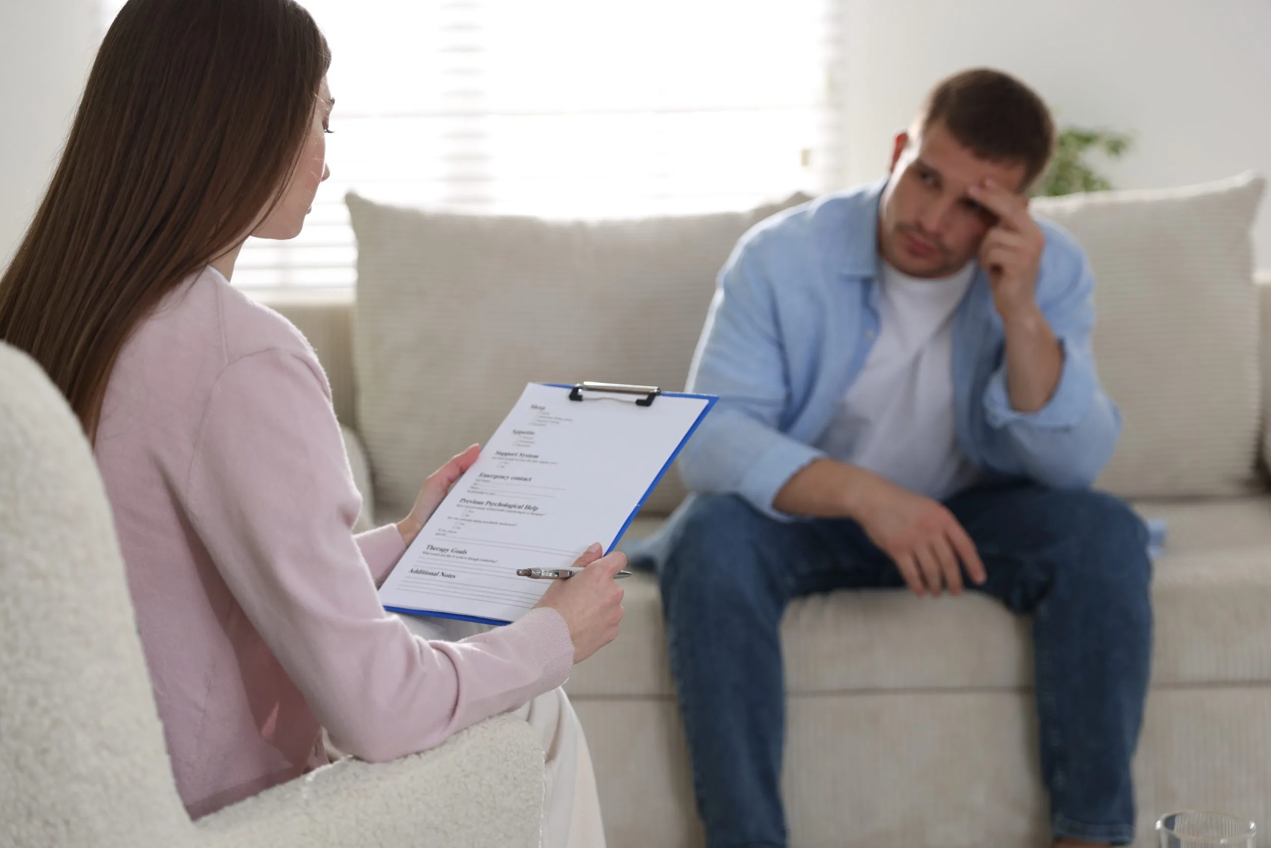 Therapist taking notes on clipboard while male patient sits on couch in therapy session