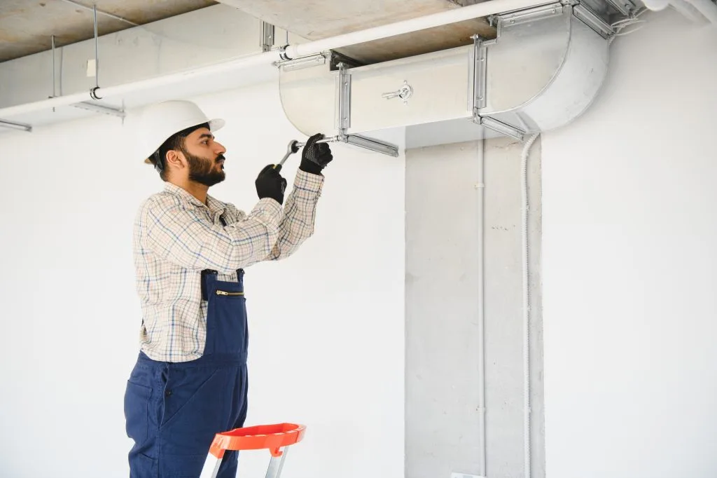 Technician in hard hat installing metal ventilation ductwork on commercial ceiling