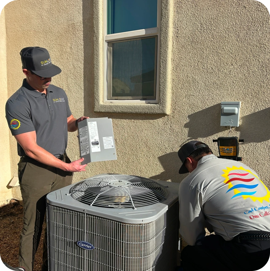 Two Sunlogix technicians installing a Carrier air conditioning unit on exterior building wall, one holding electrical panel documentation