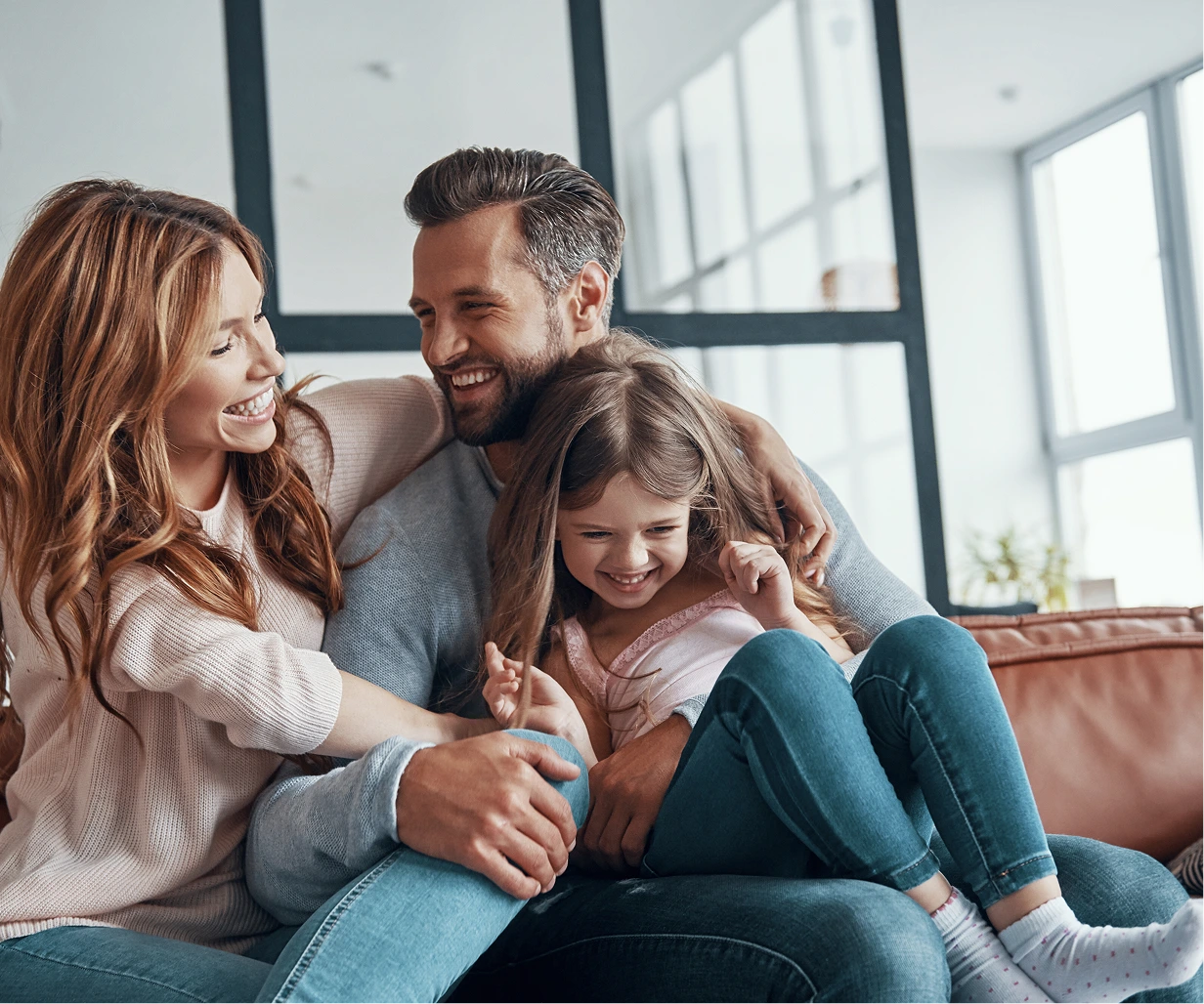 Family of four smiling together on a couch in a bright living room with large windows