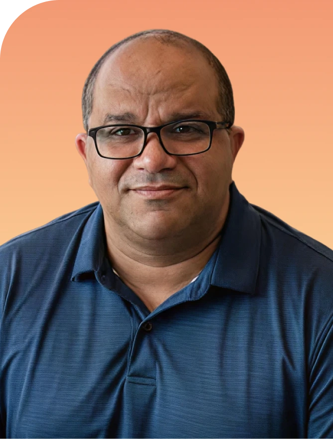 Gustavo Guzman, wearing glasses and a dark blue shirt, smiling at the camera against an orange background