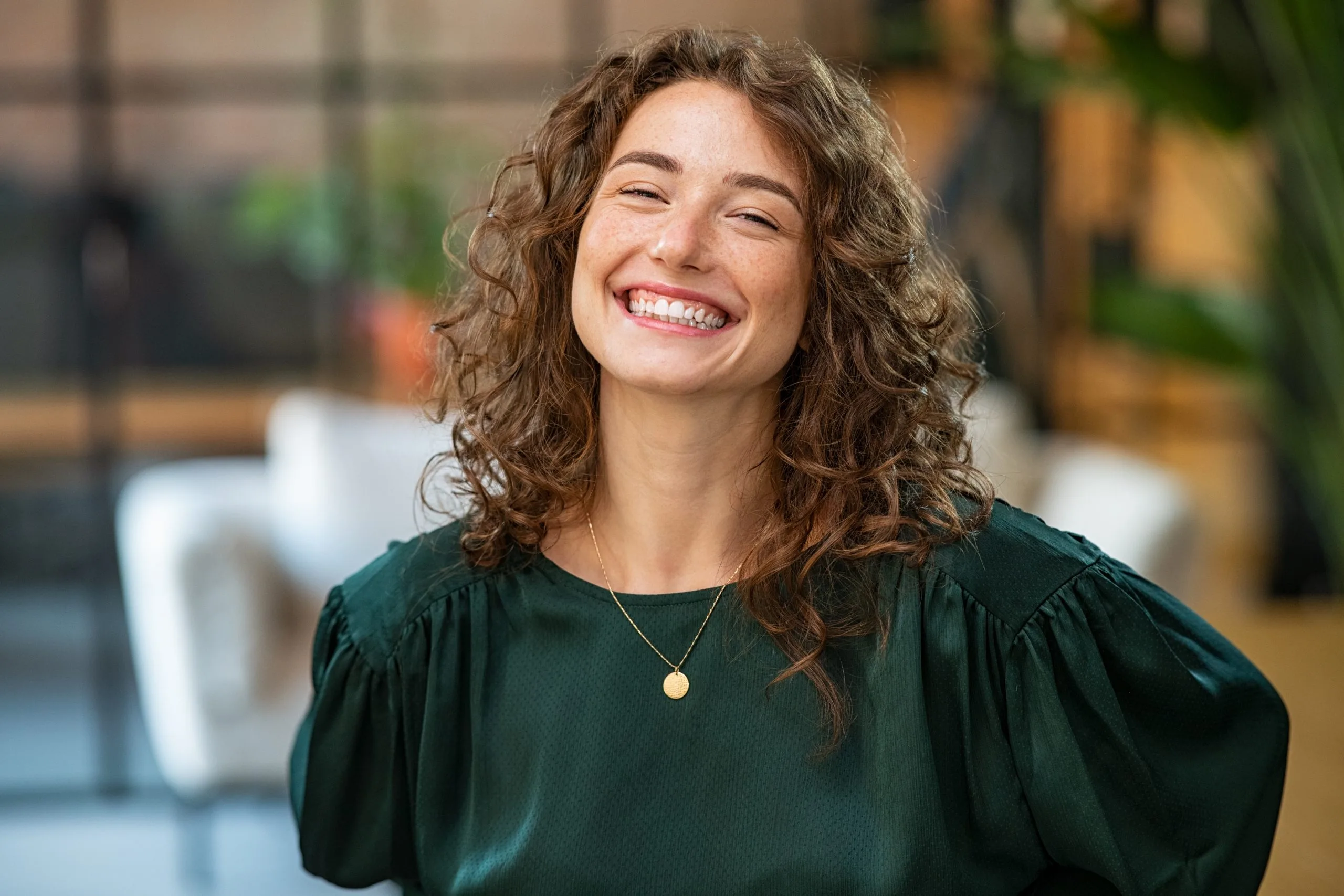 Woman with curly brown hair wearing green shirt smiling indoors with plants in background