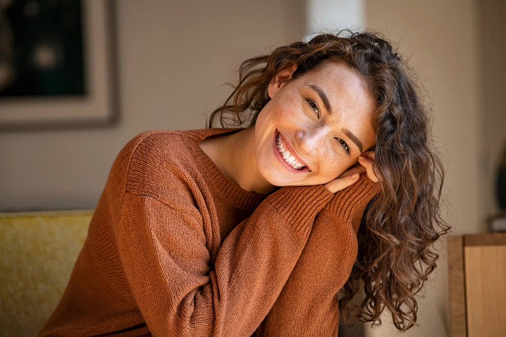 Smiling woman with wavy brown hair wearing a rust-colored sweater, hugging herself indoors