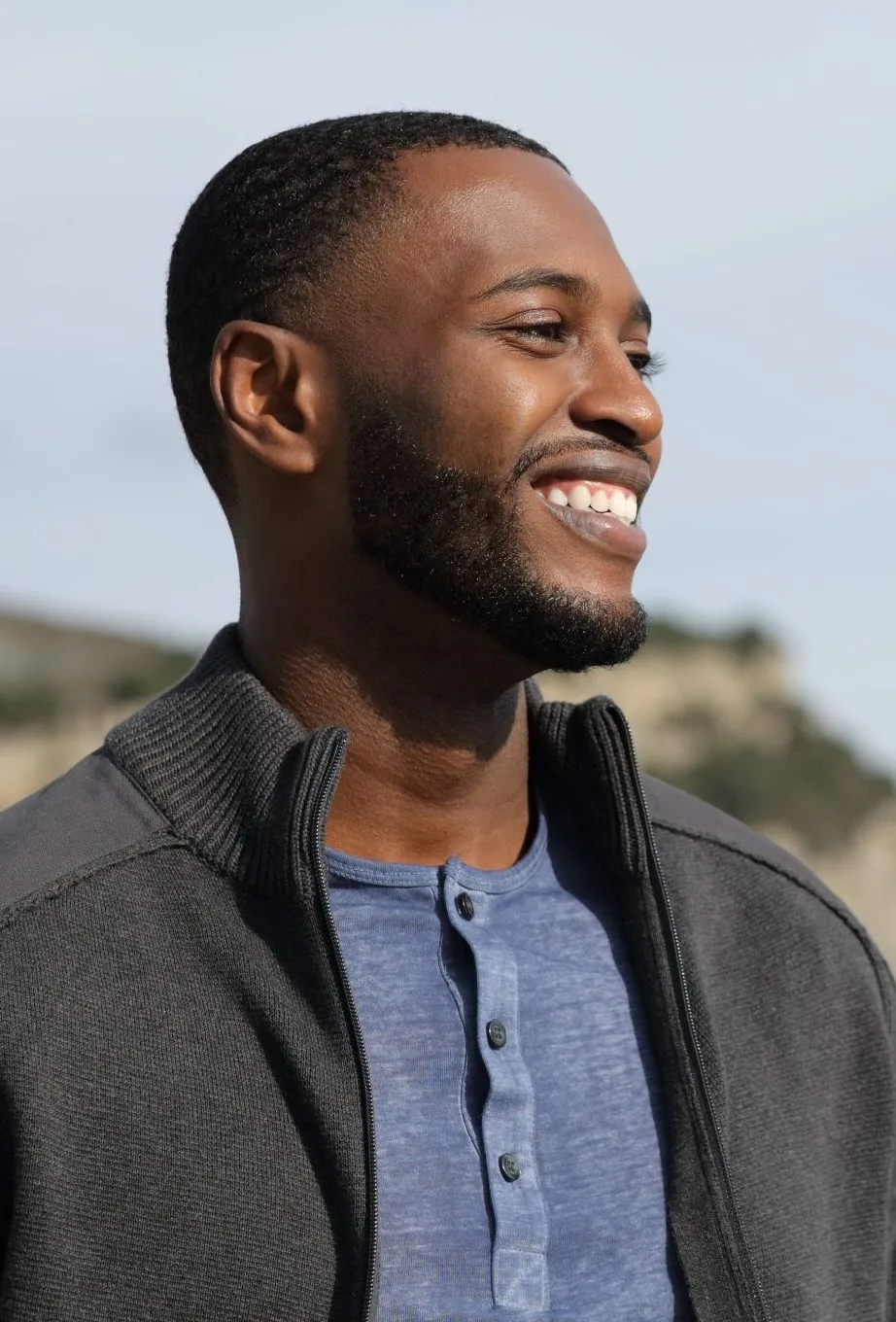 Man with short dark hair and beard wearing gray jacket over blue shirt, smiling in profile outdoors