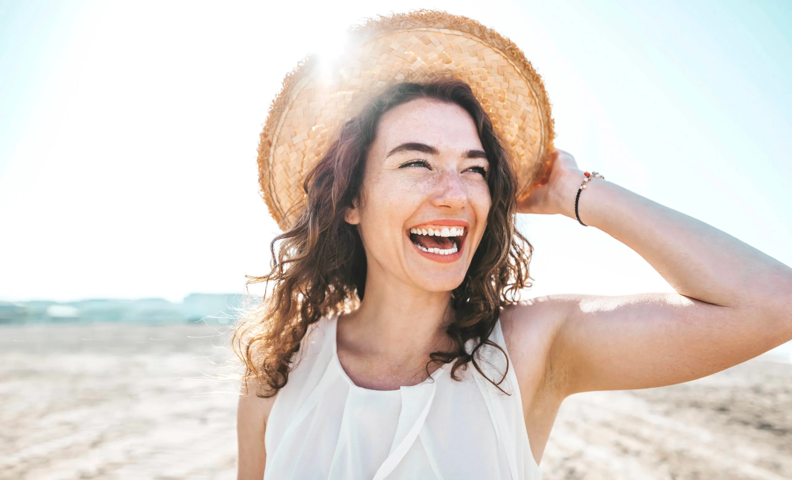 Woman wearing straw hat and white tank top laughing on beach at sunset