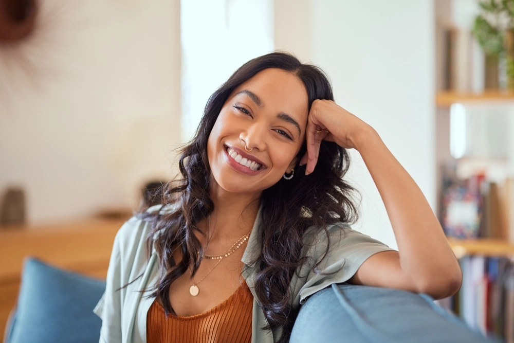 Smiling woman with long dark hair resting her head on her hand, sitting indoors on a blue chair