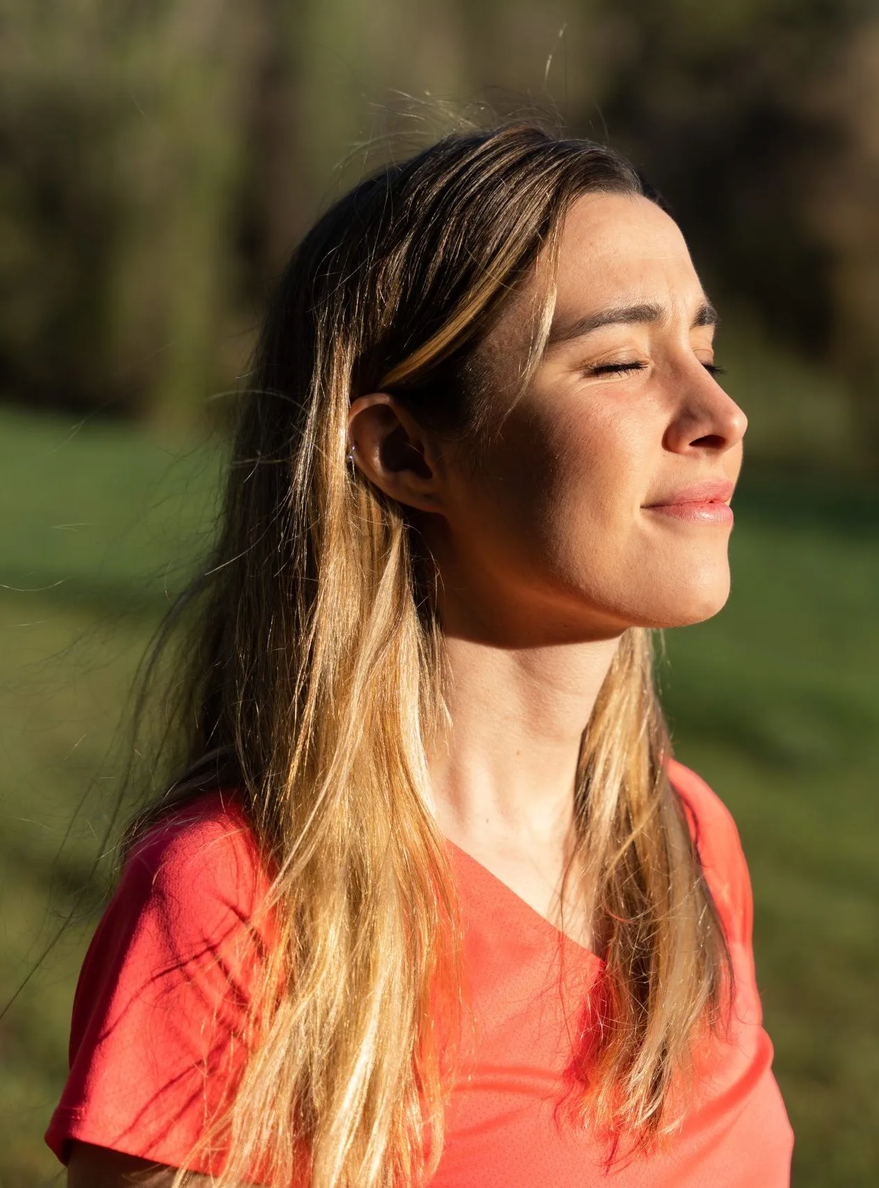 Woman with long brown hair wearing coral shirt in profile with eyes closed outdoors