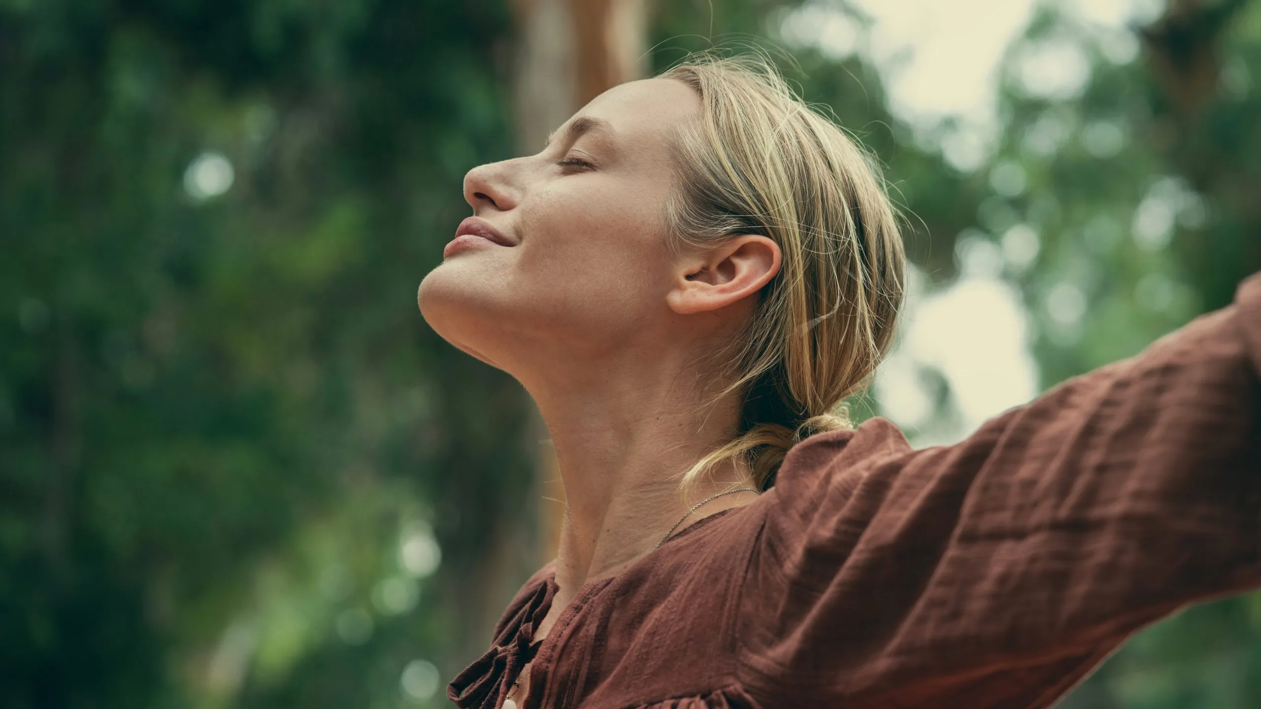 Woman in brown shirt outdoors with arms outstretched and head tilted back