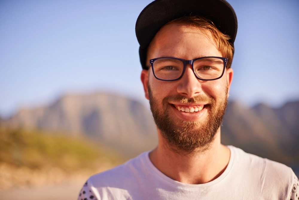 Man wearing glasses and black cap smiling outdoors with mountain landscape behind him