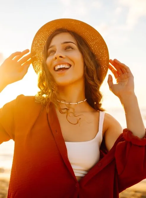 Woman in straw hat and red cardigan smiling at sunset on a beach