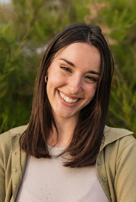 Smiling woman with long dark hair wearing a tan cardigan outdoors among green shrubs