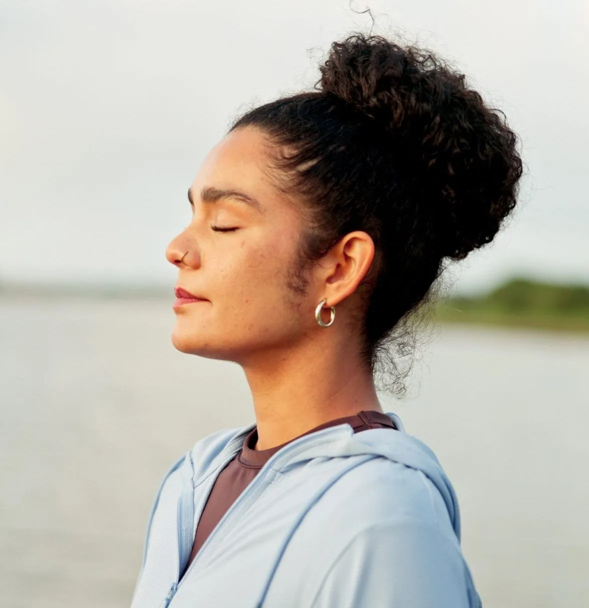 Woman with dark hair in bun, eyes closed, wearing light blue jacket against beach and sky background