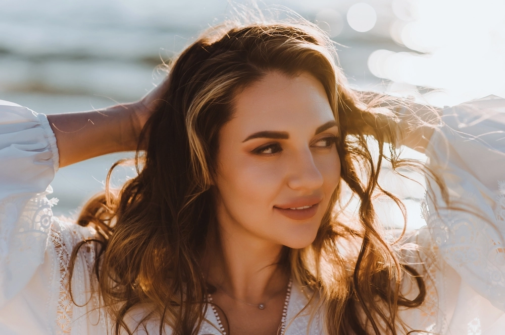 Woman with long brown hair smiling by ocean water in golden sunlight