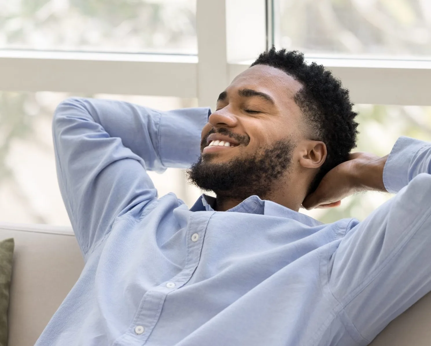 Man relaxing in chair with hands behind head, smiling by bright window