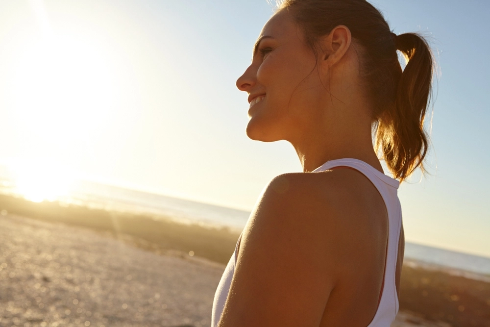 Woman in white tank top looking toward horizon at sunset on beach