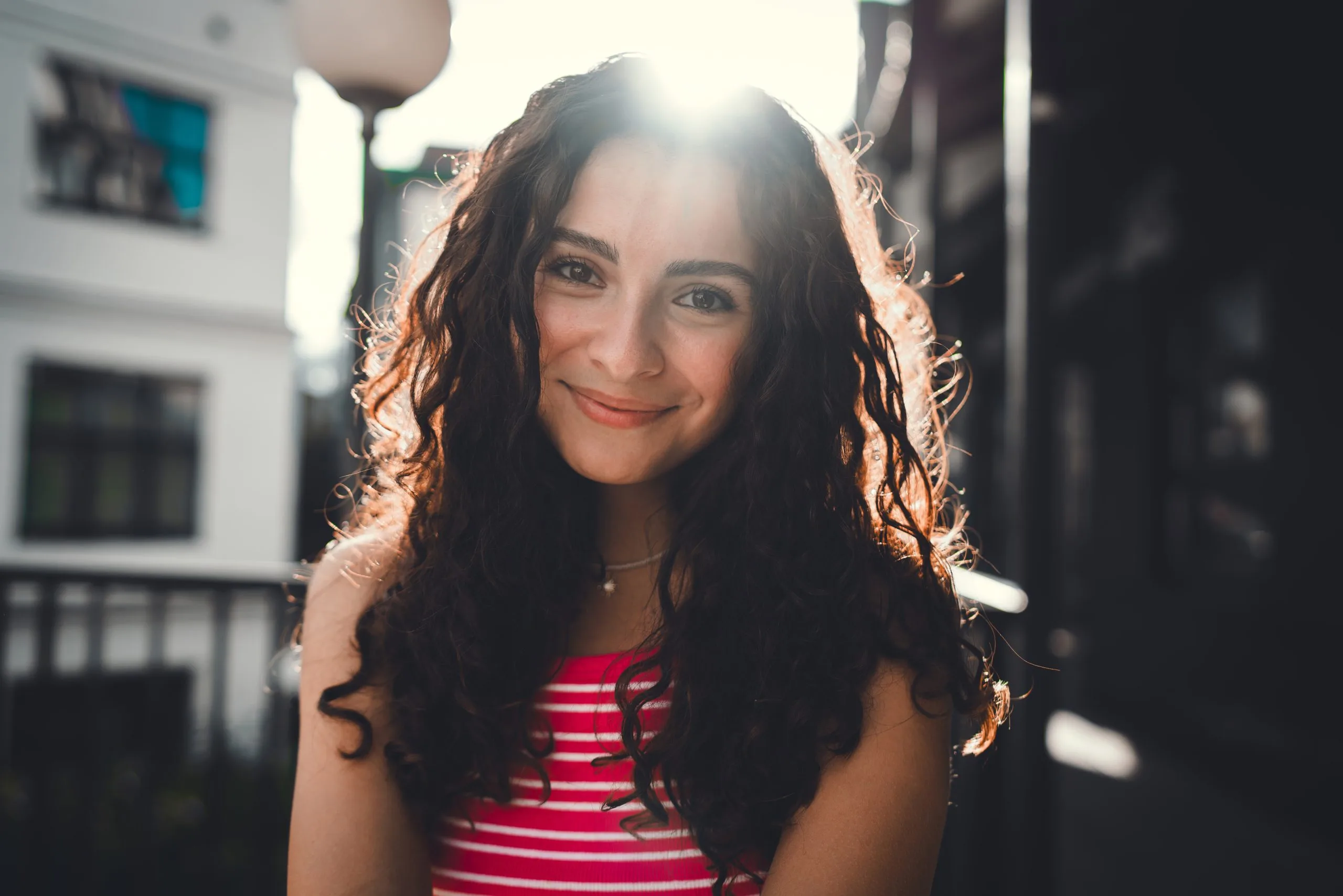 Girl with long dark curly hair wearing a red striped shirt, smiling at camera in sunlit indoor setting