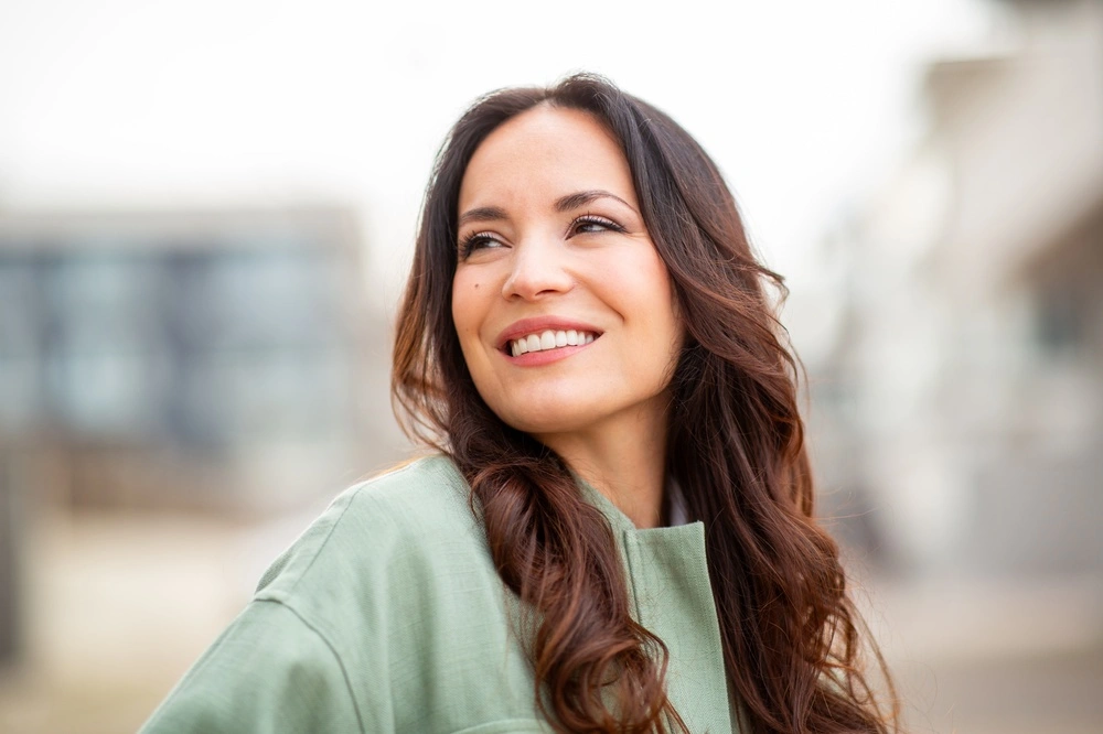 Woman with long dark hair smiling outdoors wearing a green top