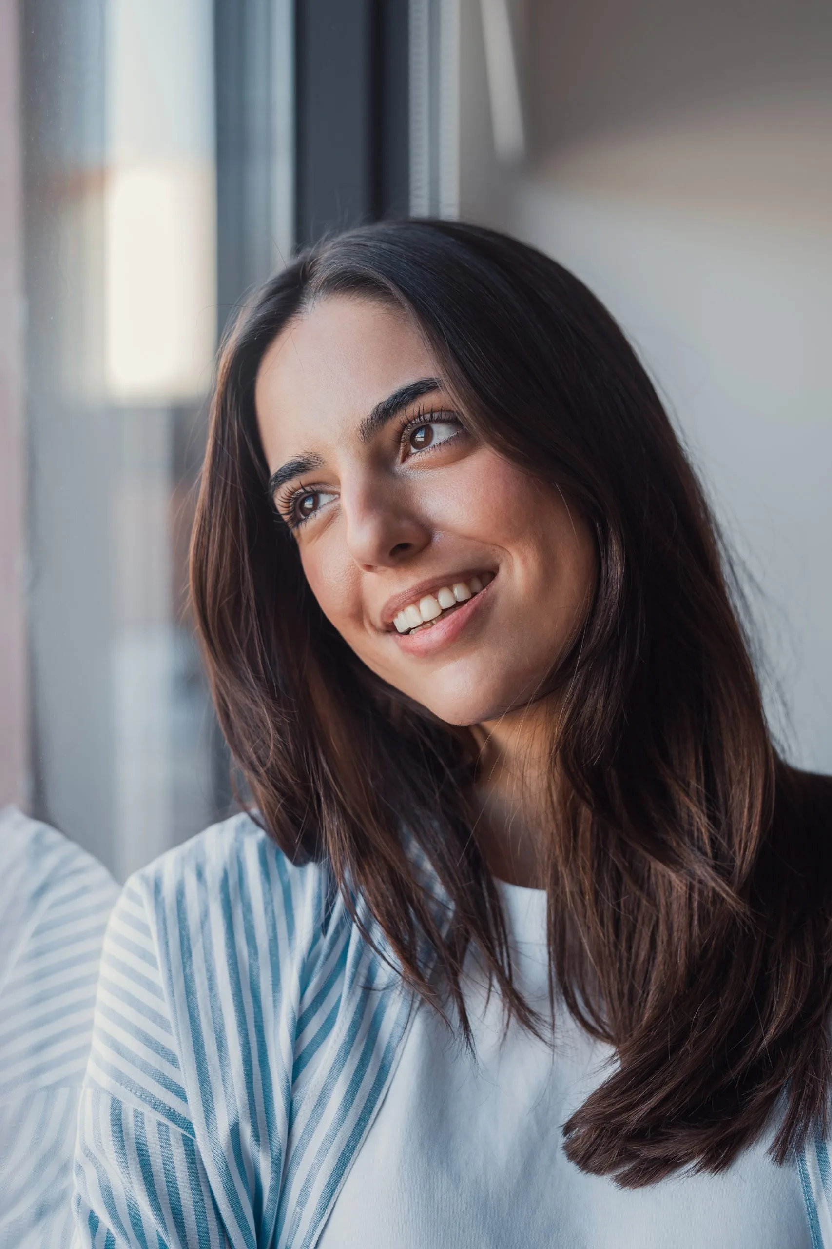 Woman with dark hair smiling by window in blue striped shirt