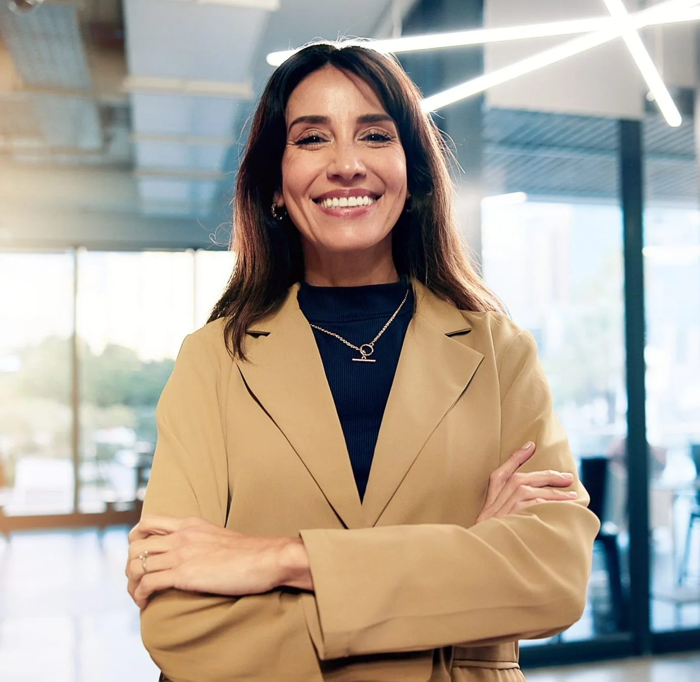 Woman in tan blazer and dark turtleneck smiling with arms crossed in modern office with skylights