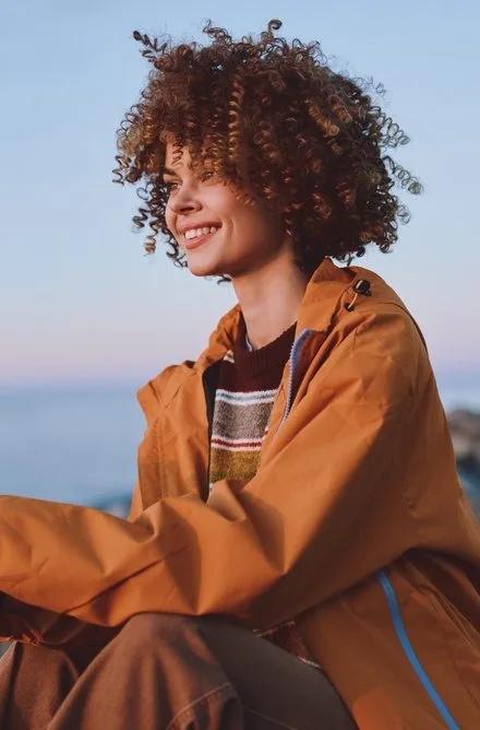 Person with curly hair sitting on rocky coastline at sunset wearing an orange jacket