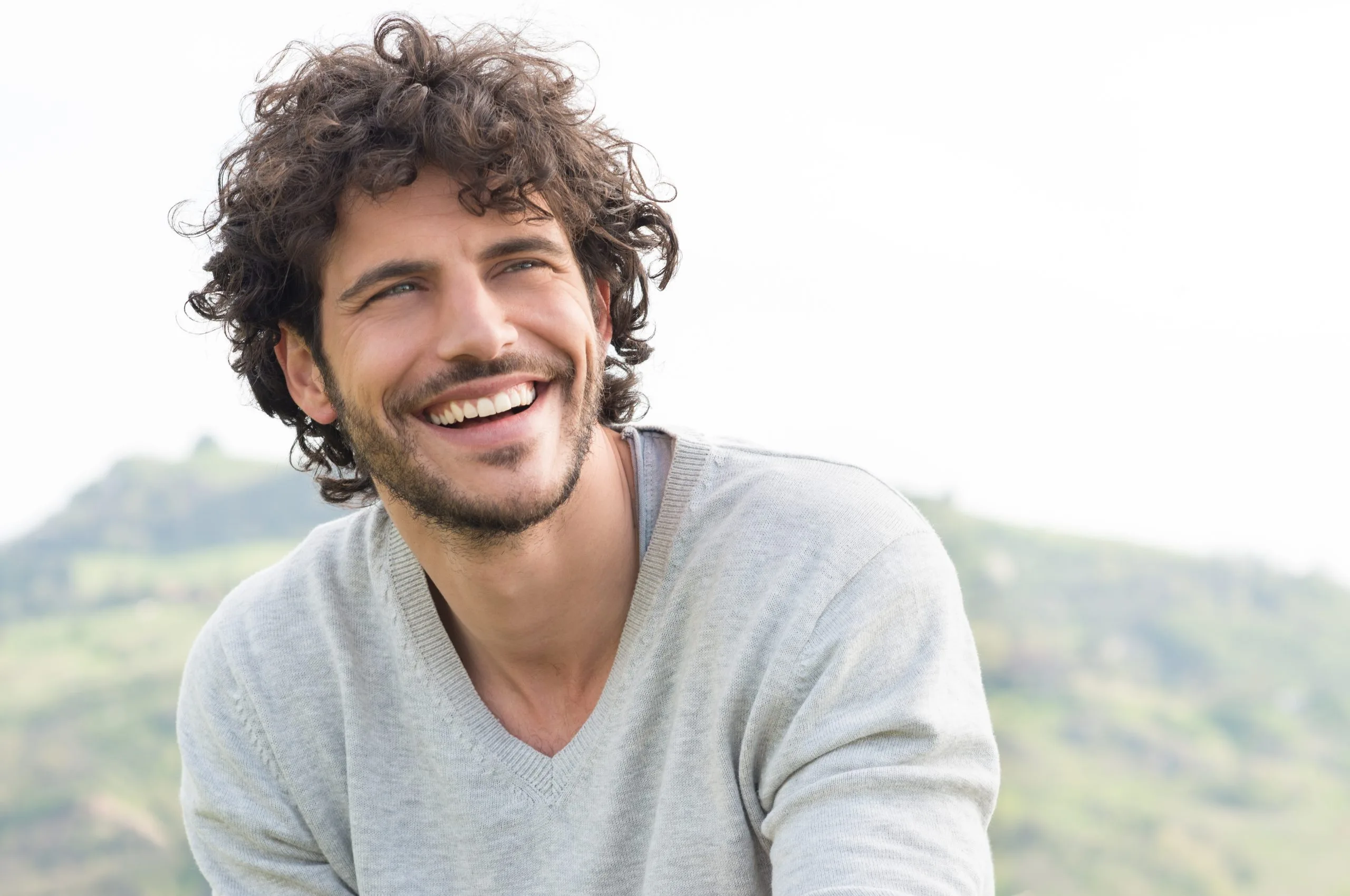 Smiling man with curly brown hair wearing a light gray v-neck shirt outdoors