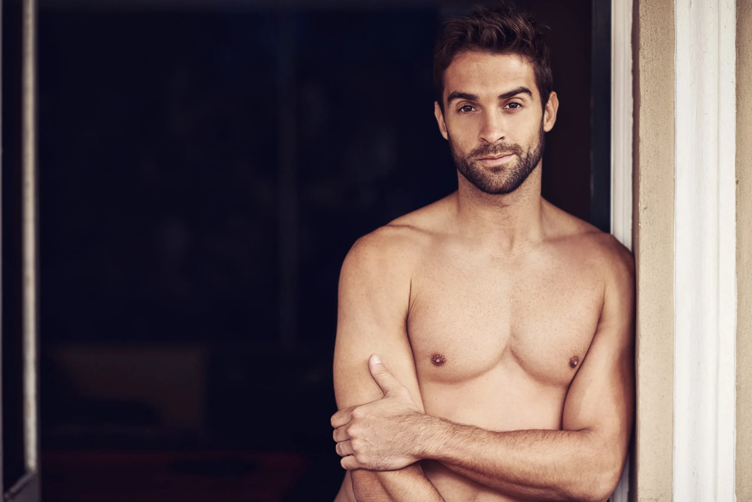 Shirtless man with crossed arms standing indoors against a dark background