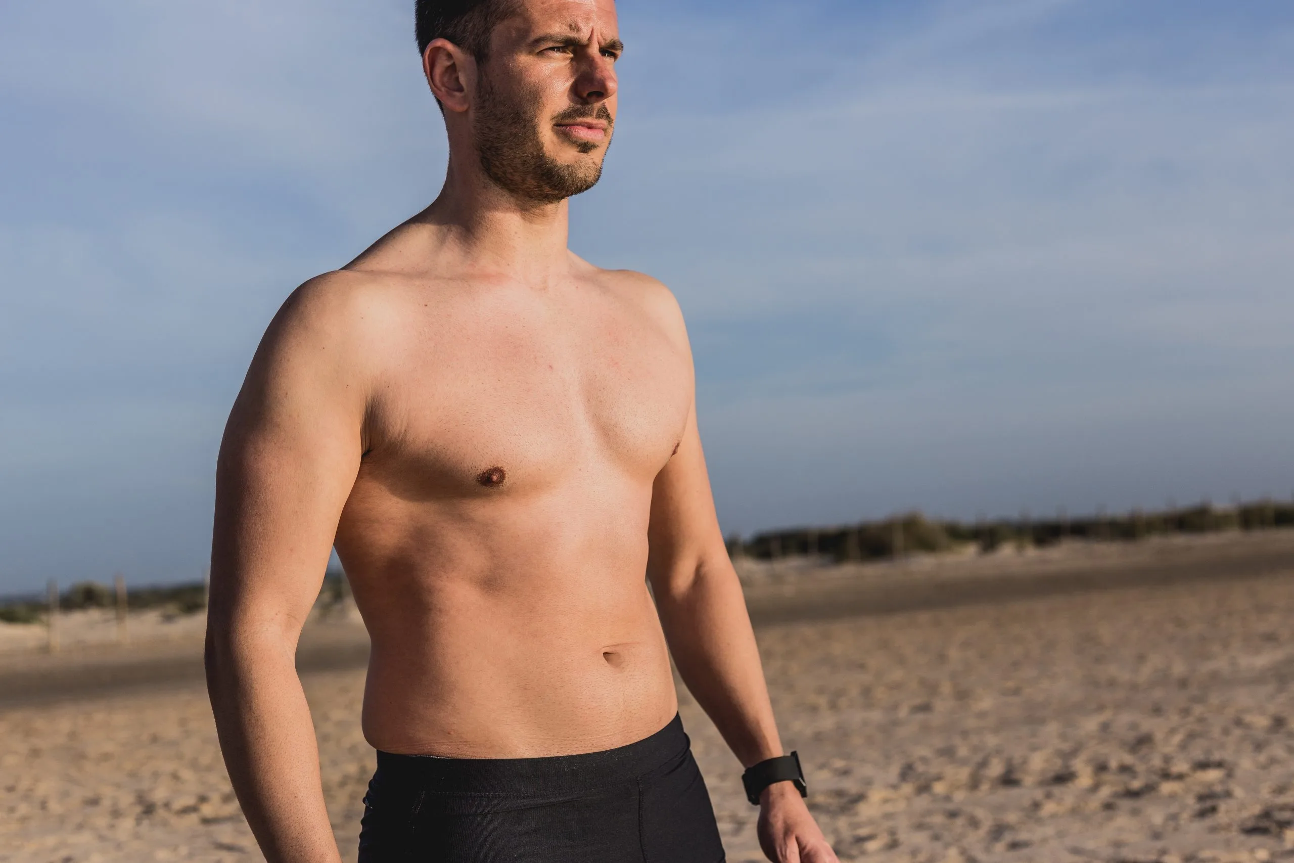Shirtless man standing on beach with sand dunes visible behind him