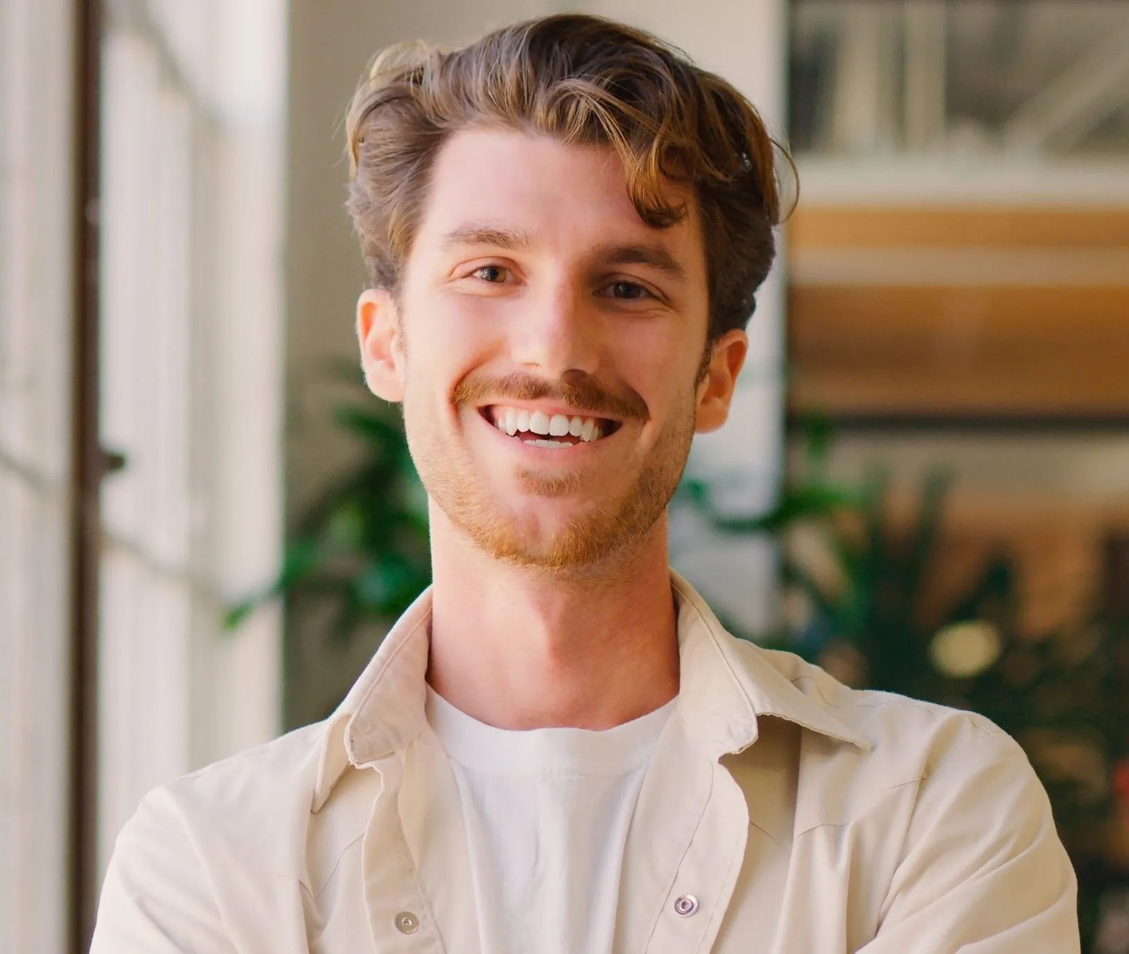 Smiling man with brown hair wearing a beige shirt over white t-shirt indoors