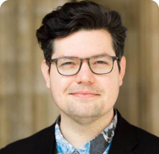 Man with dark hair wearing glasses, a black blazer, and blue patterned shirt