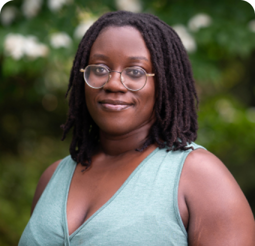 Woman with glasses wearing a light blue sleeveless top, smiling at camera with blurred green foliage background