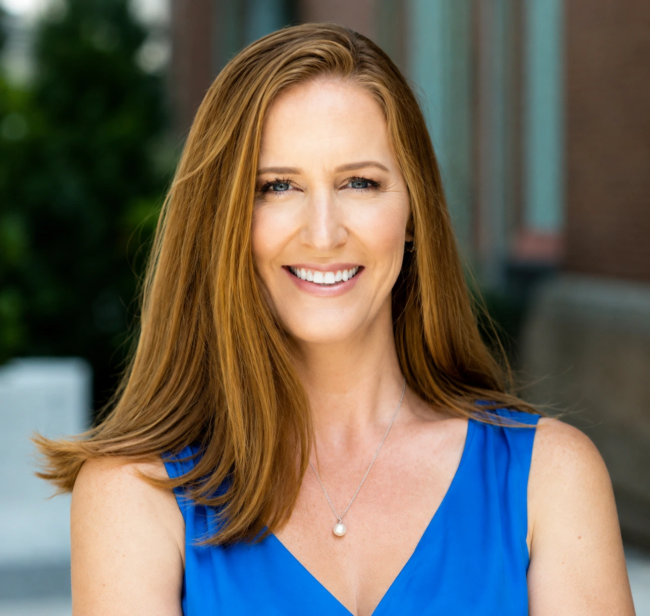 Professional headshot of a woman with long auburn hair wearing a blue sleeveless top, smiling at the camera