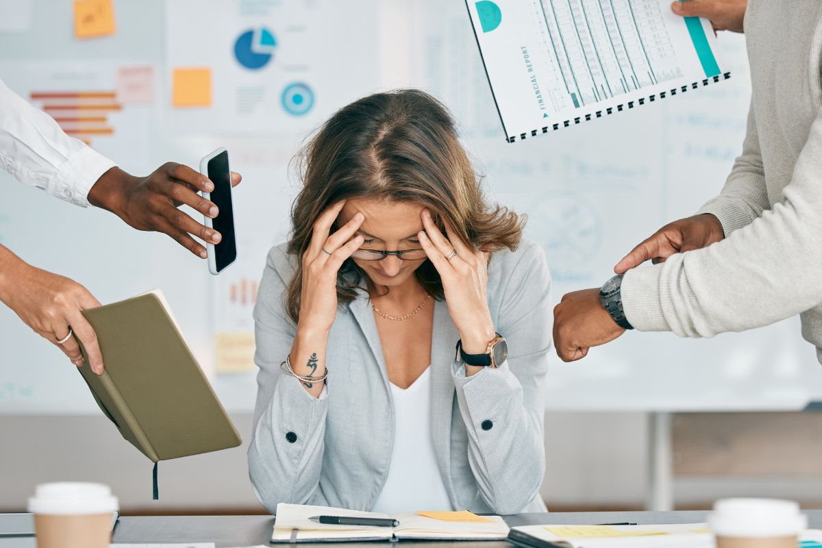 Woman at desk holding her head while surrounded by colleagues holding documents and devices, charts visible on wall behind her