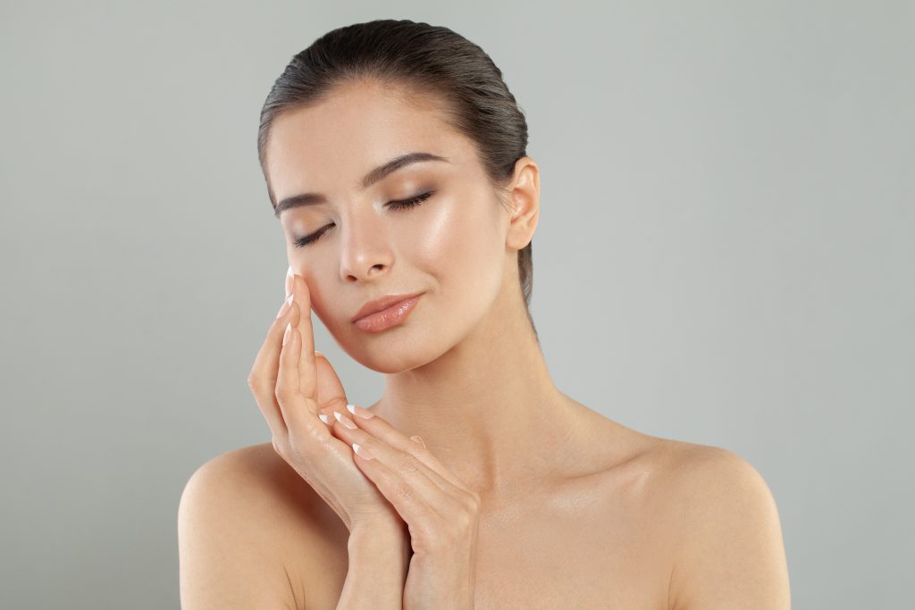 Woman with glowing skin touching her cheek with closed eyes against gray background