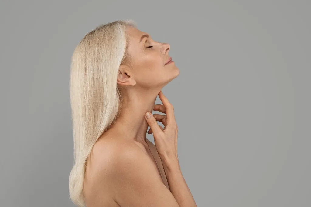 Woman with long blonde hair tilting head back and touching neck area against gray background